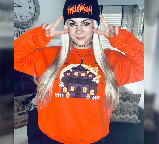 Person wearing an orange Halloween-themed sweatshirt with a haunted house design and a black beanie with orange text.